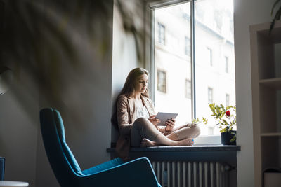 Businesswoman using tablet pc sitting on window sill at home