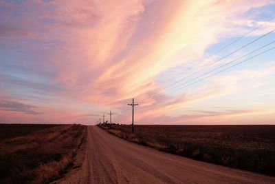 Empty dirt road by power lines against cloudy sky during sunset