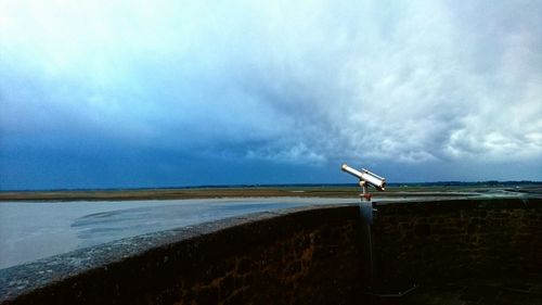 Binocular on railing by lake against cloudy sky