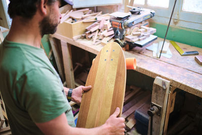 Side view of crop mature bearded male artisan with longboard against professional equipment in workshop