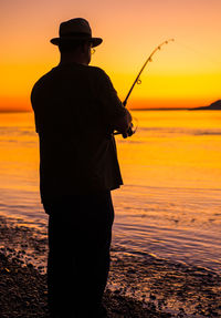 Rear view of silhouette man fishing at beach during sunset