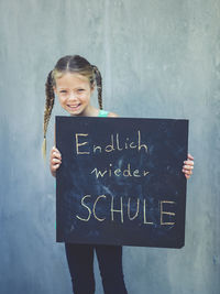 Portrait of smiling girl holding blackboard with text while standing against wall