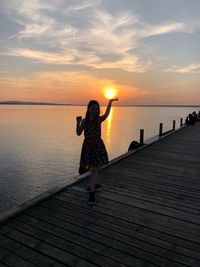 Optical illusion of girl holding sun while standing on pier over sea during sunset