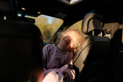 Adorable little girl sitting on black passenger seat with closed eyes in modern automobile during road trip on sunny day