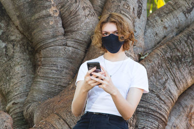 Portrait of young woman using mobile phone while standing against tree