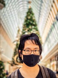 Portrait of man in eyeglasses and face mask against christmas tree and skylight inside mall.