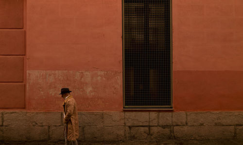 Adult man in hat and coat walking in front of red wall on street during sand storm day. madrid.