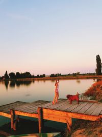 Girl standing with dog on pier over lake against sky during sunset