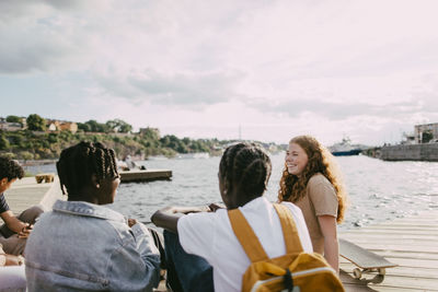 Happy teenage girl talking with male friends while sitting on pier near lake at sunny day
