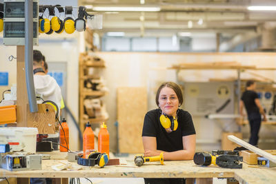 Portrait smiling young female carpentry trainee leaning on workbench with power tools at illuminated workshop