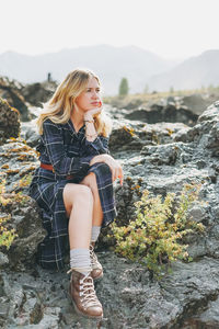 Thinking young blonde woman in blue dress sitting on stones on background of mountains