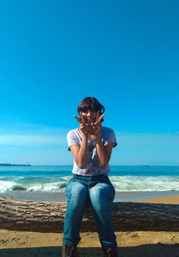 Portrait of young woman gesturing while sitting on log at beach against clear blue sky