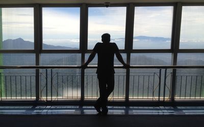 Rear view of silhouette man looking through window while standing by railing