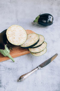 Slices of eggplant or aubergines on chopping wooden board and knife over gray background.