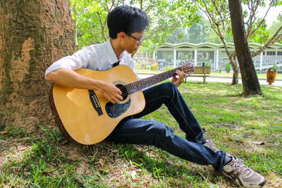 A young vietnamese man playing the guitar near a big tree in the garden