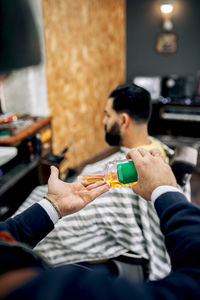 High angle of crop unrecognizable barber pouring hair oil from bottle on hand while standing near hispanic bearded male customer in traditional barbershop