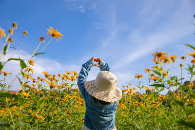 Rear view of girl making heart shape with hands while standing by flowering plants against sky