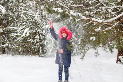 Little funny girl in red warm hat shakes the snow from a branch outside on nature winter snowy 