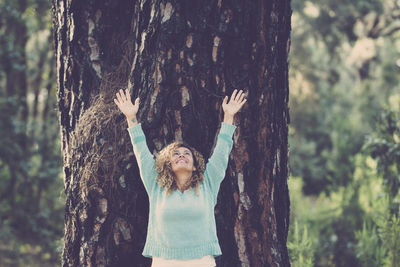 Woman with arms raised standing by tree trunk in forest