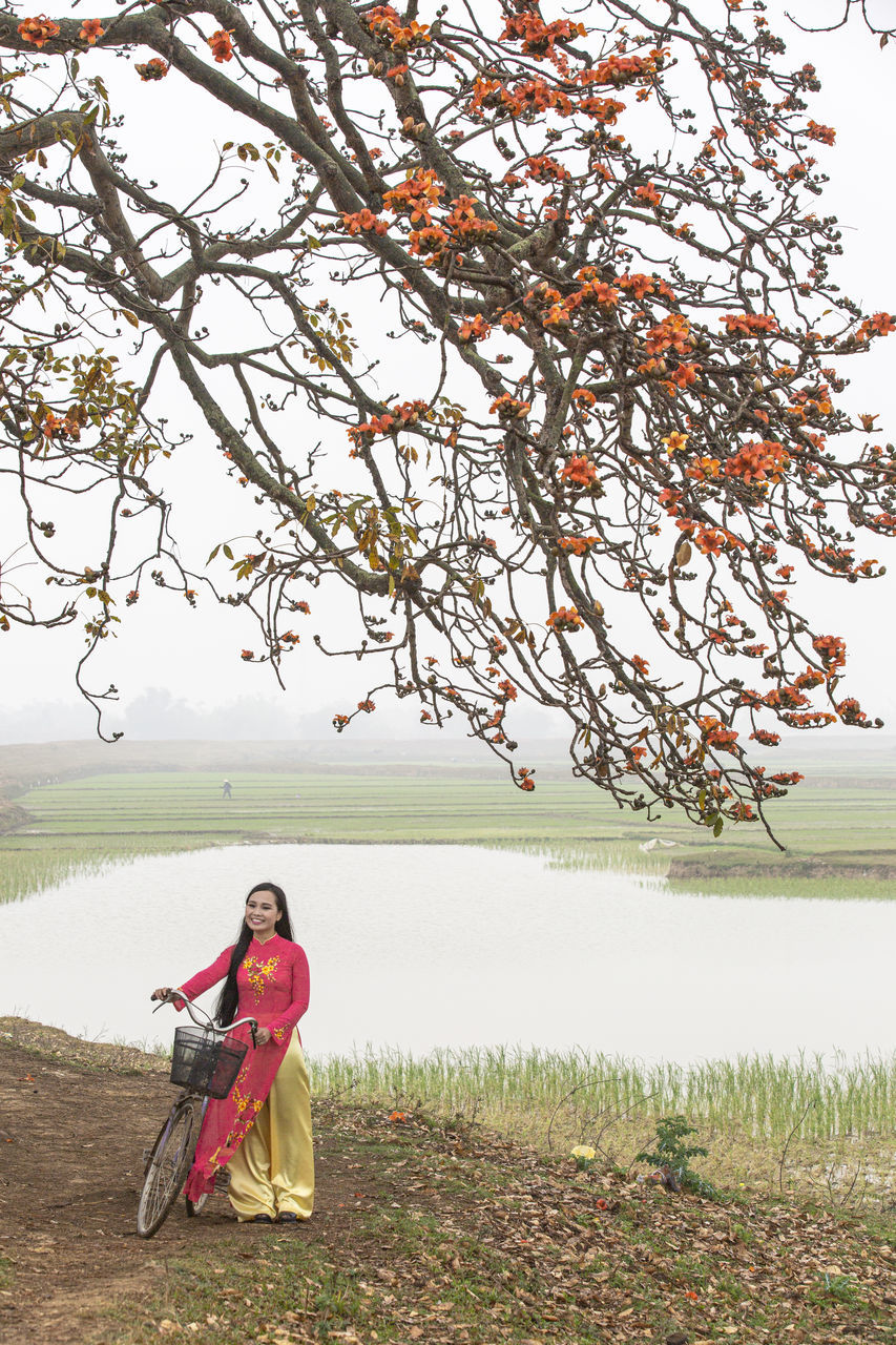 plant, nature, flower, tree, one person, water, adult, full length, lake, beauty in nature, tranquility, sky, women, clothing, autumn, landscape, sitting, tranquil scene, day, outdoors, rural scene, grass, environment, leisure activity, lifestyles, land, red, scenics - nature, relaxation, traditional clothing