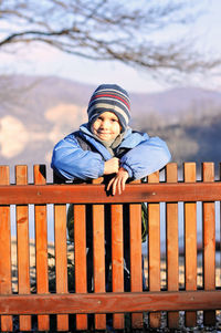 Portrait of boy leaning on wooden fence against mountains