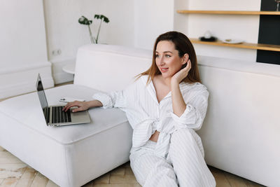 A millennial woman in white pajamas looks into a laptop and works online using technology