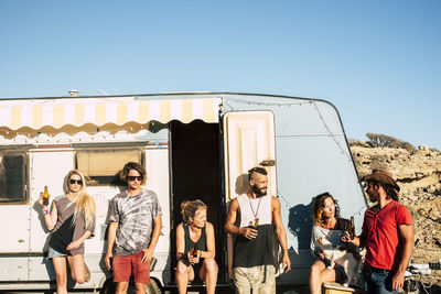 Friends holding beer bottles standing by motor home against sky