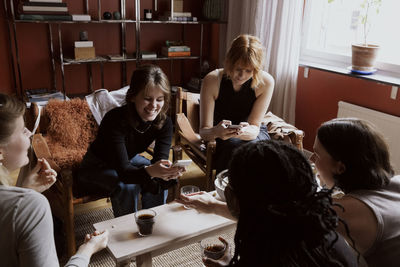 Smiling women using smart phones while celebrating with female friends in apartment