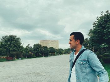 Young man looking away while standing by trees against sky