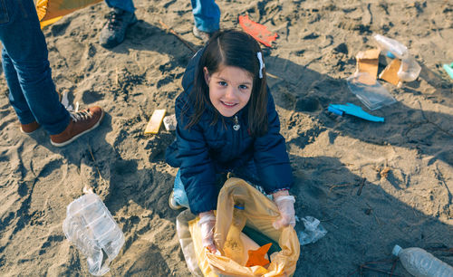 High angle portrait of girl holding garbage in plastic bag while couching at beach