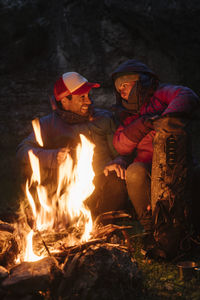 Cyclists arranging halt and warming at fireplace preparing sausages on stick at green meadow at night