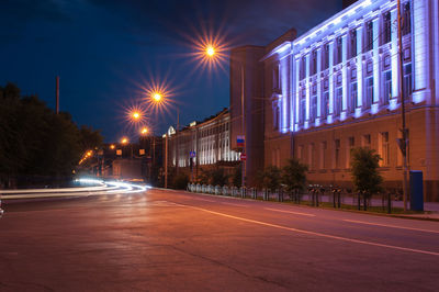 Illuminated road by buildings against sky at night