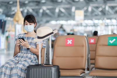 Woman wearing mask using smart phone sitting at airport