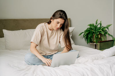 A young brunette woman in a t-shirt and jeans sitting on the bed works on a laptop