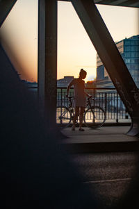 Woman standing by bicycle on bridge in city during sunset