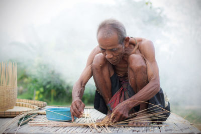 Full length of shirtless senior man making wicker basket