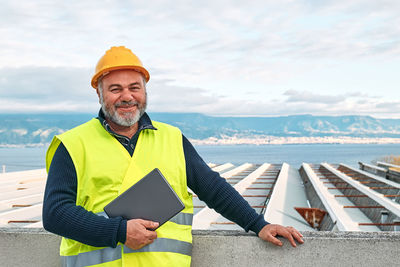 Portrait of smiling middle aged supervisor in hardhat and safety vest with tablet on building site.