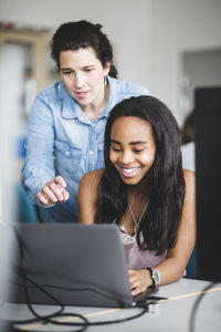 Confident female teacher assisting smiling high school student using laptop at desk in classroom