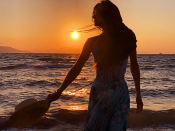 Rear view of woman standing at beach against sky during sunset