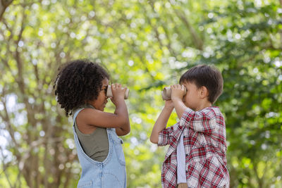 Side view of sibling looking through binoculars against trees
