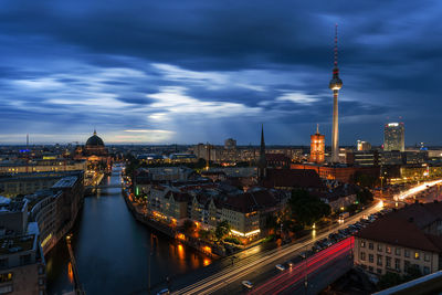 Distant view of illuminated fernsehturm against cloudy sky at dusk