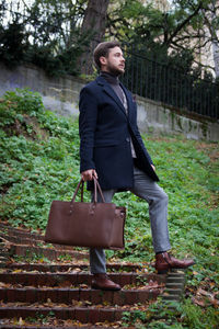 Full length of thoughtful businessman holding bag while standing outdoors