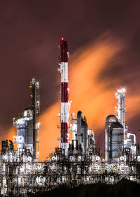 Illuminated factory by buildings against sky at night