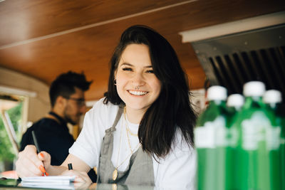 Portrait of happy female owner writing order in food truck while male coworker working in background