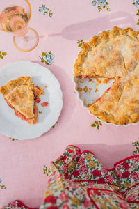 Single slice of rhubarb pie on white plate, pink tablecloth with blue roses, rose wine, floral apron