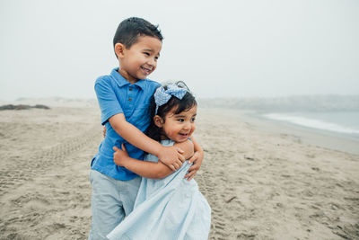 Young siblings smile and embrace at beach looking toward the waves