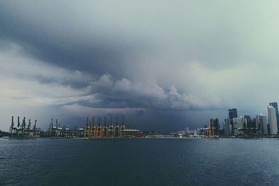 Factories and buildings in city by sea against cloudy sky