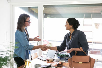 Female coworkers giving handshake while standing at table in home office