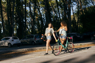 Female friends walking on street holding skate board and bicycle in vacations