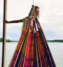 Portrait of woman seen through multi colored hammock by river against sky